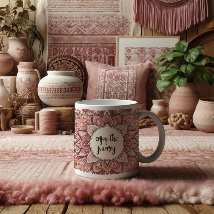 White ceramic mug with a pink and brown mandala design and the phrase 'Enjoy the Journey' in the center, placed in a cozy bohemian-style room with pink and earthy-toned decor, including patterned cushions, pottery, and indoor plants.