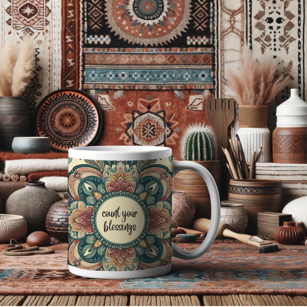 White ceramic mug with a detailed green, red, and gold mandala design and the phrase 'Count Your Blessings' in the center, placed in a cozy bohemian-style room with earthy-toned decor, including patterned rugs, pottery, woven baskets, and indoor plants.